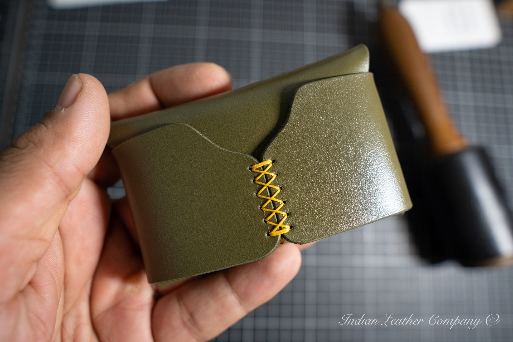 Hand holding a handmade corsair green leather wallet with yellow stitching on a checkered background
