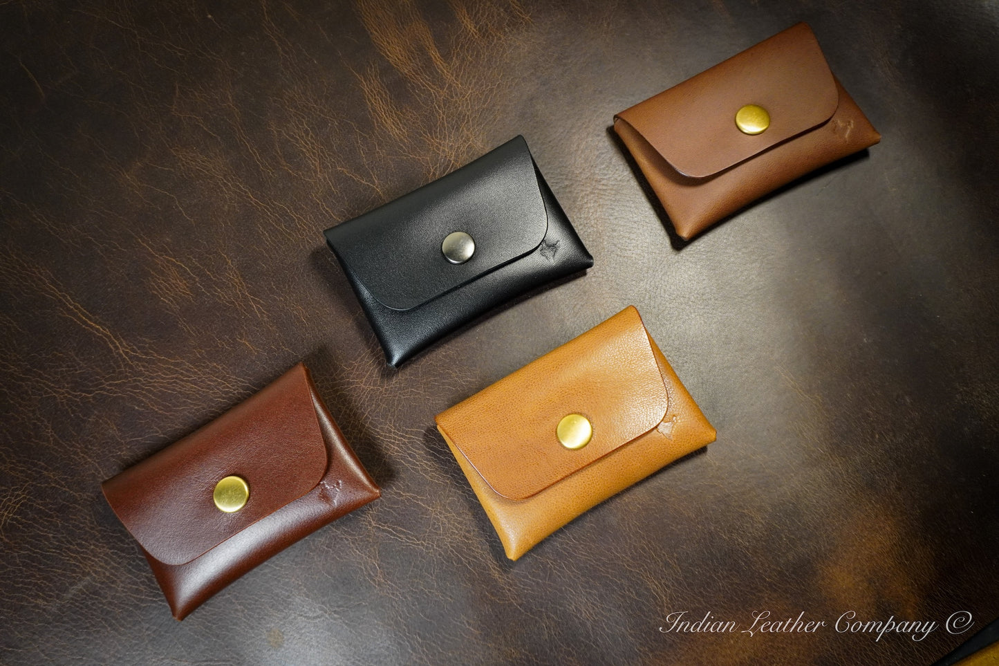 Four leather wallets in black, brown, and gold on a dark leather surface with 'Indian Leather Company' branding.