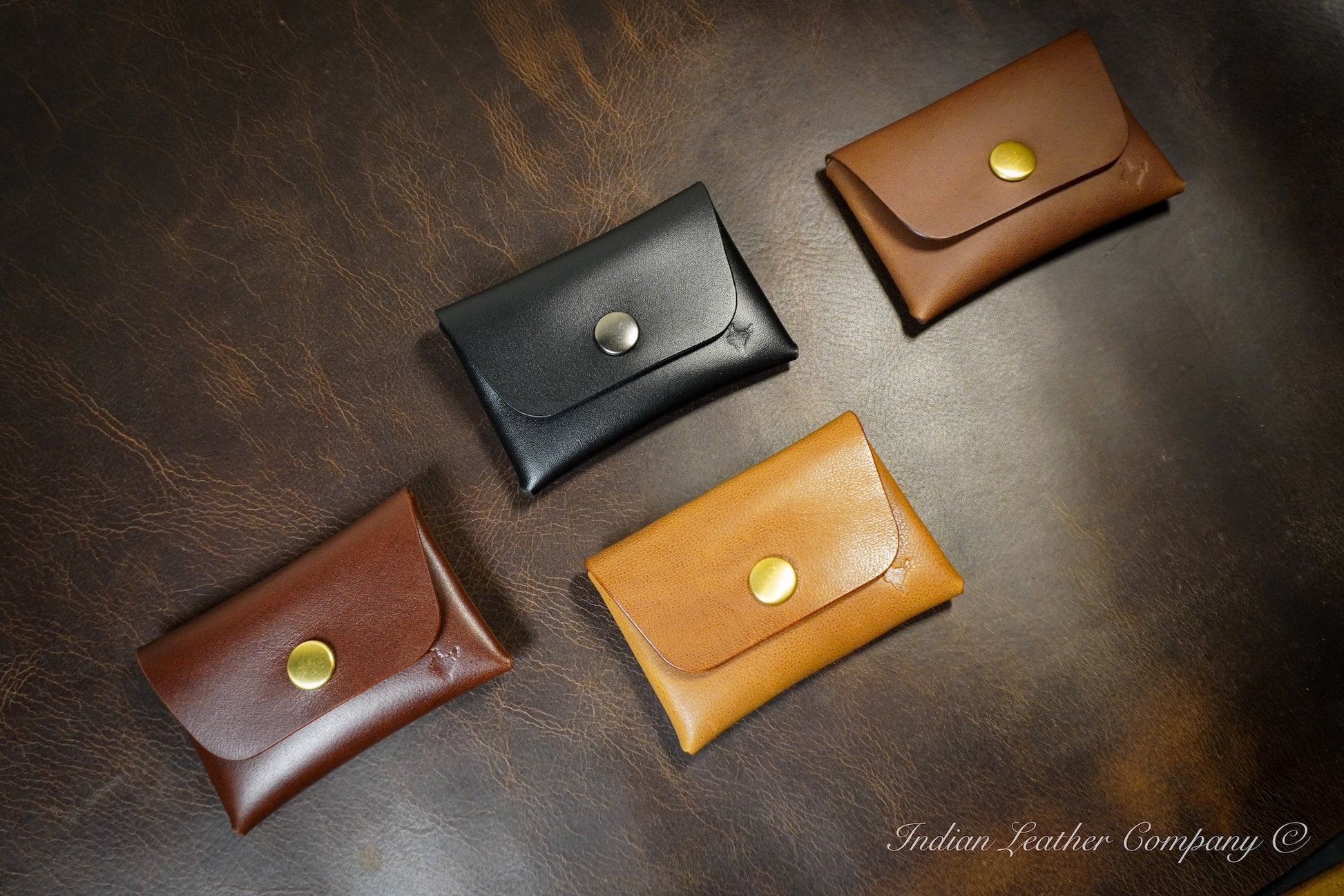 Four leather wallets in black, brown, and gold on a dark leather surface with 'Indian Leather Company' branding.