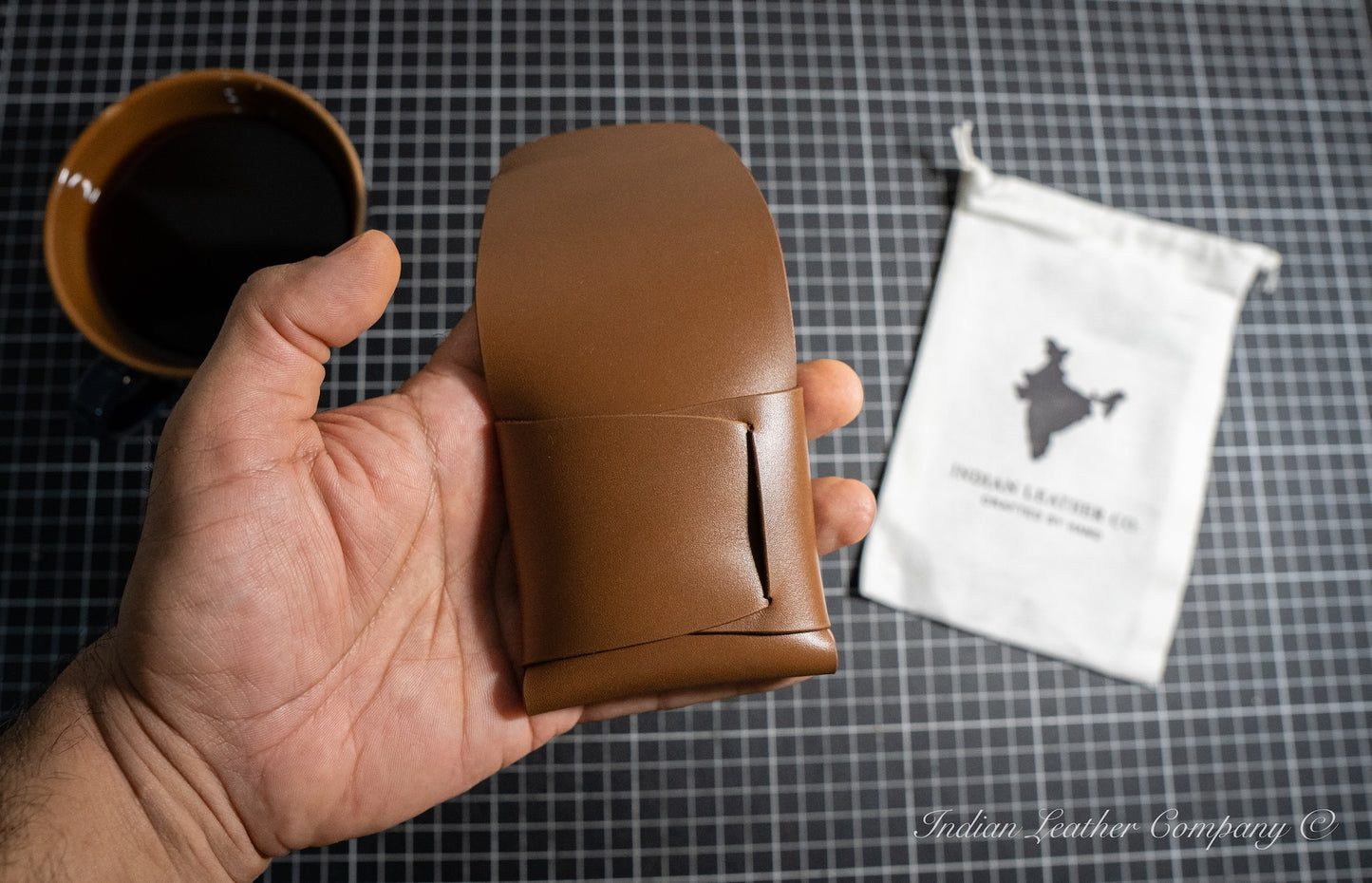 Back side of the Brown full grain wallet in hand with a coffee cup in the background and Indian Leather company white bag