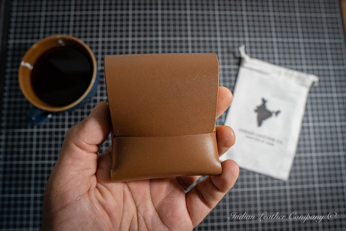 Hand holding a brown leather wallet with a cup of coffee and a package in the background.