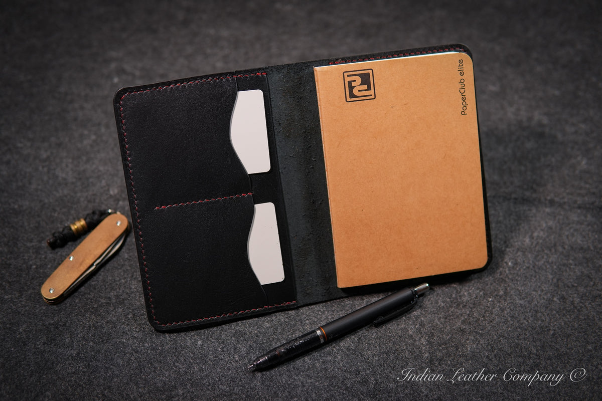 A black full grain veg tanned leather notebook cover with red stitching, shown open on a dark textured surface. The cover includes two card slots, a notebook insert, a black pencil, and a small folding knife.