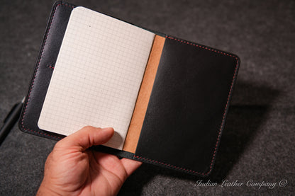 An open black leather notebook cover held by a hand, showing a white paper notebook and red stitching on a dark textured background.