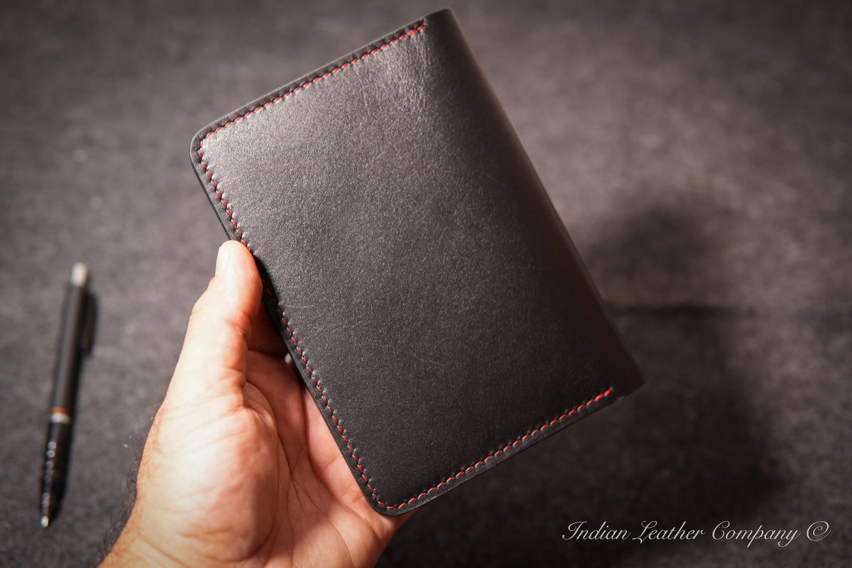 A hand holding a black leather journal cover with red contrast stitching, set against a dark textured background with a pen on the left