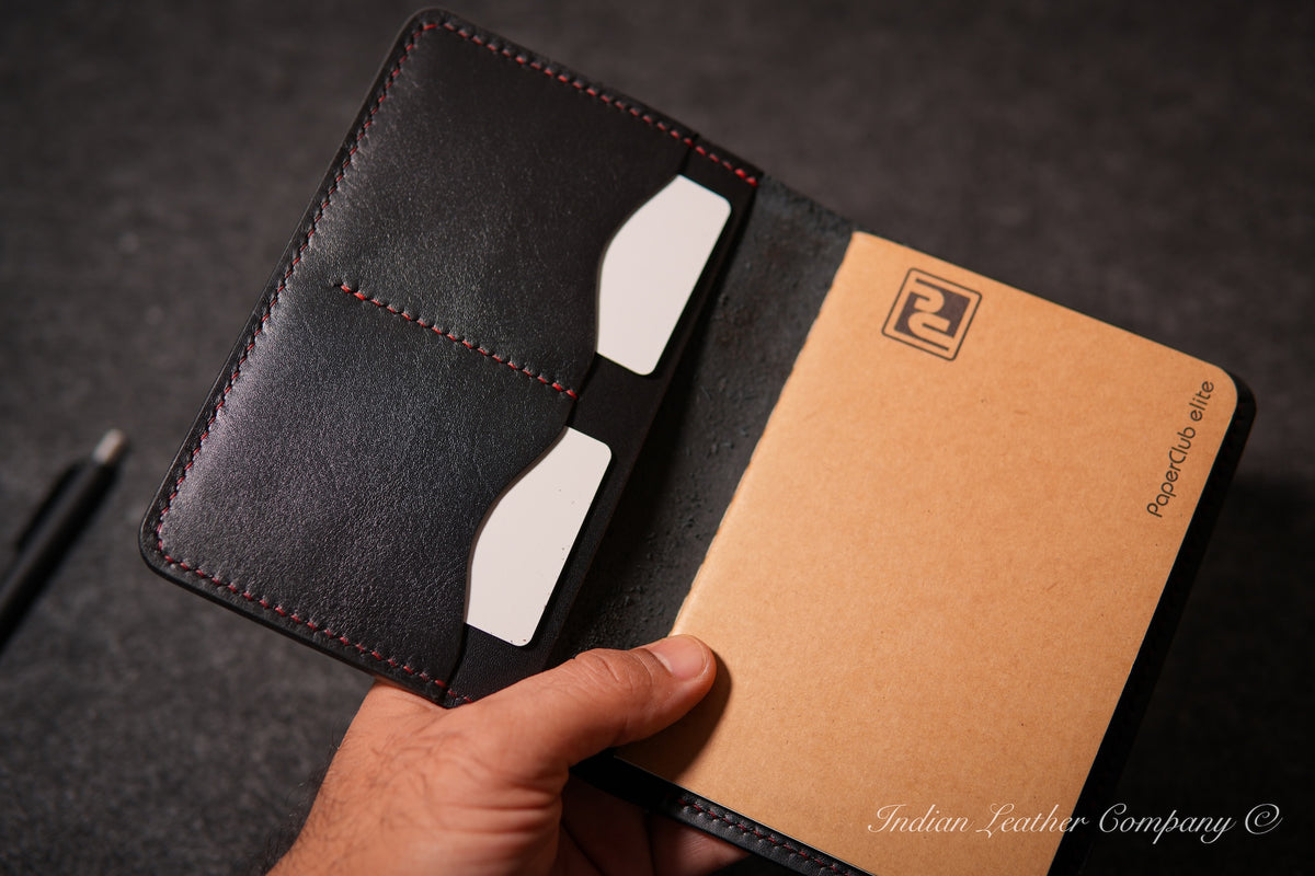 A hand holding an open black leather notebook cover with red stitching, showing a tan kraft-paper style interior page and card slots with white cards. 